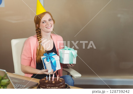 Woman sitting at home office desk holding wrapped gifts with chocolate cake and laptop, copy space 128739242