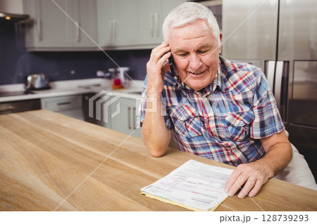 Senior man talking on smartphone, reviewing papers at wooden island in home kitchen, copy space 128739293