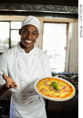 African American male chef smiling and presenting freshly baked pizza in commercial kitchen African American male chef smiling and presenting freshly baked pizza in commercial kitchen 128739296