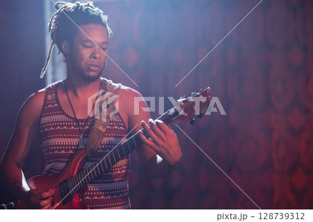 Male musician playing electric guitar on dim stage under colored lights, with decorated strap Male musician playing electric guitar on dim stage under colored lights, with decorated strap 128739312