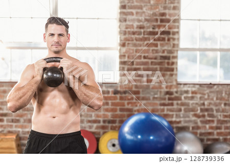 Man lifting kettlebell at chest level in industrial gym, featuring weight plates and exercise balls Man lifting kettlebell at chest level in industrial gym, featuring weight plates and exercise balls 128739336