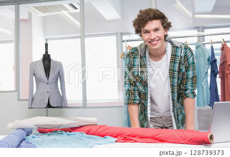 Smiling male fashion designer leaning on worktable in studio, with measuring tape and fabric rolls 128739373