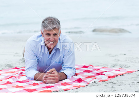 Smiling senior man lying on sandy shoreline at quiet beach, with red and white picnic blanket 128739374