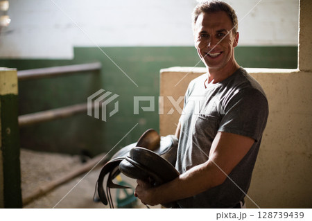 Smiling mature man holding dark leather saddle in barn aisle, with stable background, copy space Smiling mature man holding dark leather saddle in barn aisle, with stable background, copy space 128739439