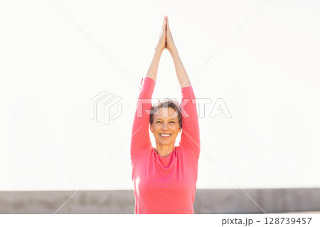 Holding arms overhead woman practicing yoga on rooftop terrace, with concrete barrier Holding arms overhead woman practicing yoga on rooftop terrace, with concrete barrier 128739457