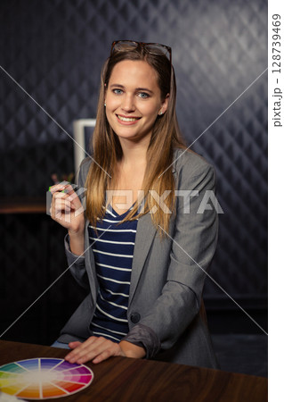 Smiling woman holding green pen near face at design studio desk, with color wheel and eyeglasses Smiling woman holding green pen near face at design studio desk, with color wheel and eyeglasses 128739469