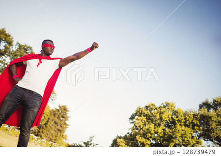 African American man standing heroically in park field, with red cape and wristbands, copy space African American man standing heroically in park field, with red cape and wristbands, copy space 128739479