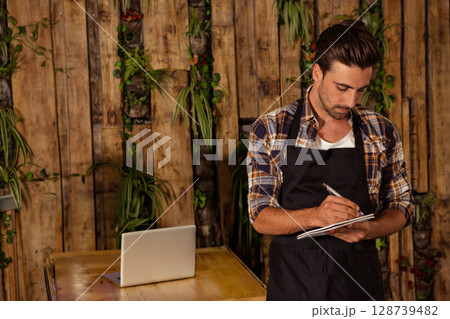 Male barista writing notes on notepad at wooden table in cafe, with laptop, plants, copy space 128739482