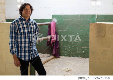 Standing man examining metal railings inside stable, with purple jacket, wood shavings, copy space 128739500