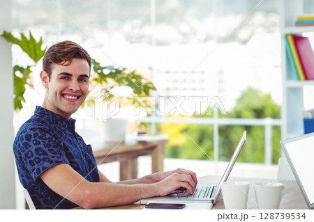 Smiling man typing on silver laptop at modern office, with coffee mugs and smartphone, copy space Smiling man typing on silver laptop at modern office, with coffee mugs and smartphone, copy space 128739534