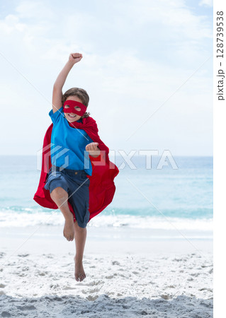Leaping child playing superhero on white sandy beach, wearing red mask, cape and blue clothes 128739538