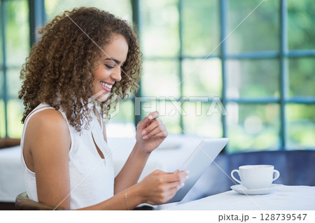 Smiling woman reading white tablet at cafe table, with white cup and saucer, copy space 128739547