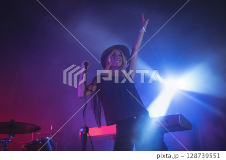 Singing woman artist holding mic stand on stage under red-purple lights, with drum kit and keyboard Singing woman artist holding mic stand on stage under red-purple lights, with drum kit and keyboard 128739551