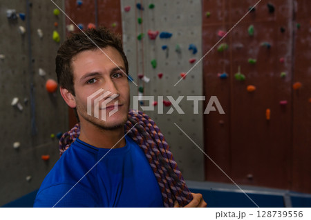 Standing man wearing blue athletic shirt at climbing gym, with rope and colorful holds, copy space Standing man wearing blue athletic shirt at climbing gym, with rope and colorful holds, copy space 128739556