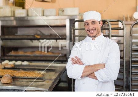 Smiling mid-adult male chef standing in bakery kitchen, with loaves and steel table, copy space Smiling mid-adult male chef standing in bakery kitchen, with loaves and steel table, copy space 128739561