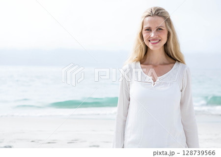 Smiling woman standing on sandy beach near calm turquoise water, wearing white blouse 128739566