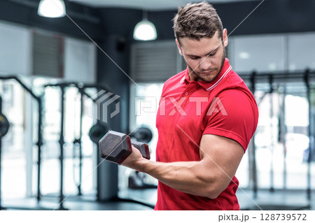 Lifting man performing bicep curl at gym, with hexagonal dumbbell and weight racks, copy space 128739572