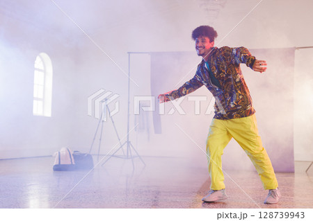 Man dancing energetically in hazy studio, with tripod, backdrop stand and duffel bag, copy space 128739943