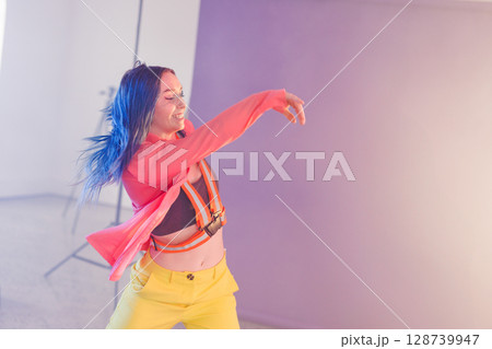 Non-binary dancer dancing in indoor photography studio, wearing orange harness with lighting stand 128739947