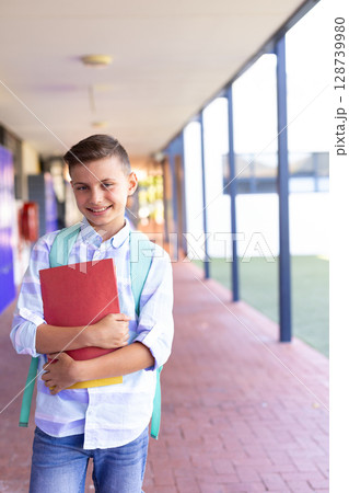 School-age boy walking in school corridor, carrying red binder yellow notebook and green backpack 128739980