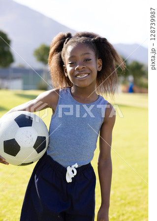 African American child holding soccer ball on grassy sports field, smiling under clear sky African American child holding soccer ball on grassy sports field, smiling under clear sky 128739987
