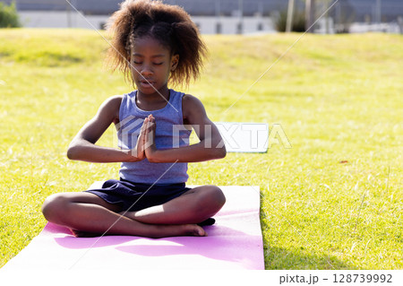 African American girl holding prayer pose on pink yoga mat in grassy park field, practicing yoga African American girl holding prayer pose on pink yoga mat in grassy park field, practicing yoga 128739992