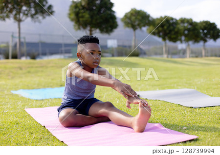 African American boy child performing forward leg stretch on grassy field, with pink yoga mat 128739994