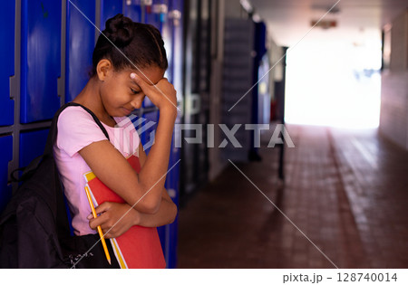 African American student standing in school hallway, with red binder and pencils, looking worried 128740014