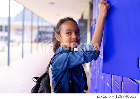 Female child student reaching up to open locker in covered school corridor, carrying black backpack 128740015