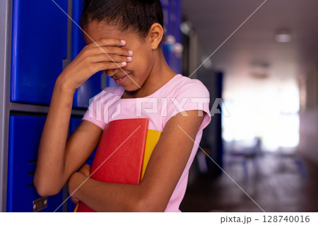 African American schoolgirl standing by blue lockers in hallway, with notebook and shy expression 128740016
