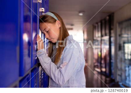 Teenage girl leaning against blue metal locker in school hallway, wearing denim jacket and headband Teenage girl leaning against blue metal locker in school hallway, wearing denim jacket and headband 128740019