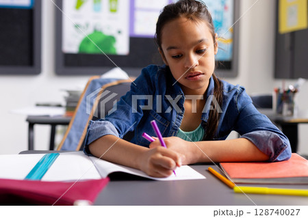 Child girl writing with purple pen in notebook at elementary classroom desk with binder, copy space 128740027
