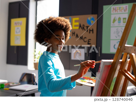 Child boy painting on easel in art classroom with paintbrush, palette and Earth Chance poster Child boy painting on easel in art classroom with paintbrush, palette and Earth Chance poster 128740040