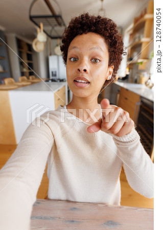 non-binary person in 20s taking selfie and pointing at camera in home kitchen, featuring pot rack 128740054