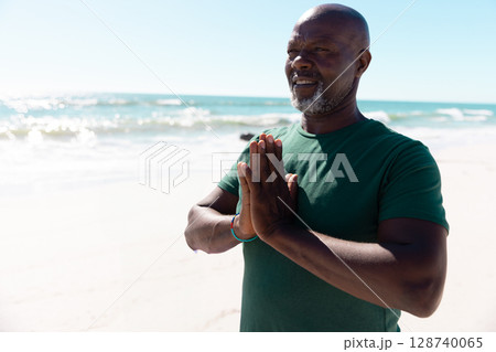 Senior African American man praying on beach water's edge, wearing colorful wristband, copy space 128740065