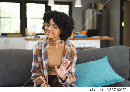 Teenage girl wearing eyeglasses sitting on grey couch in open living room waving with teal pillow 128740072