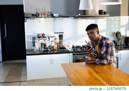Teenage boy leaning on wooden island counter in kitchen, using smartphone, copy space 128740079