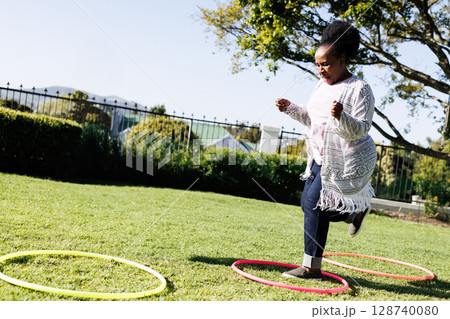 African American woman jumping into pink hoop in sunlit backyard with colorful hoops, copy space African American woman jumping into pink hoop in sunlit backyard with colorful hoops, copy space 128740080