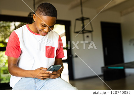 Black teenage boy sitting on bench by glass doors in modern lounge, using smartphone, copy space 128740083