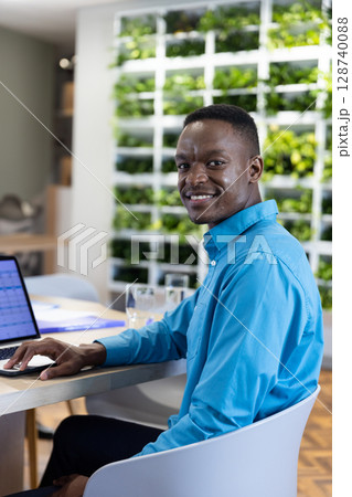 African American man sitting at desk in modern office, using silver laptop with water glasses African American man sitting at desk in modern office, using silver laptop with water glasses 128740088