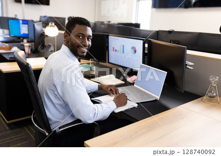 Young African American man writing in notebook smiling at camera at office desk, with laptop Young African American man writing in notebook smiling at camera at office desk, with laptop 128740092