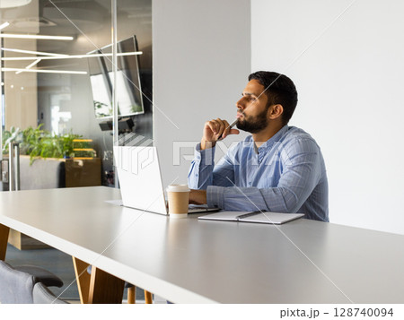 Man sitting at table in open-plan office, with laptop, coffee cup and notebook, copy space 128740094