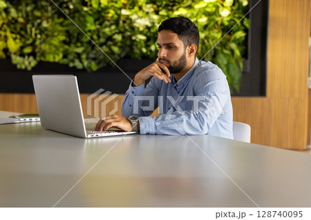 Young man working on silver laptop at white table in office, with plant wall, copy space 128740095