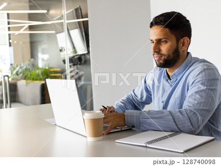 Man typing on silver laptop at conference table in modern office, with coffee cup and notebook Man typing on silver laptop at conference table in modern office, with coffee cup and notebook 128740098