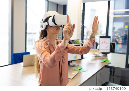 Mid adult woman standing at desk in modern office, wearing VR headset and manipulating interface 128740100