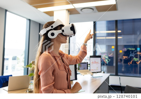 Mid-adult woman wearing VR headset reaching toward virtual interface at office desk, with monitors 128740101