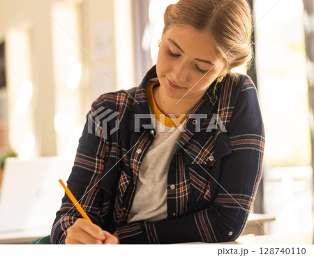 Woman writing on paper at table in sunlit classroom, with yellow pencil on tabletop 128740110