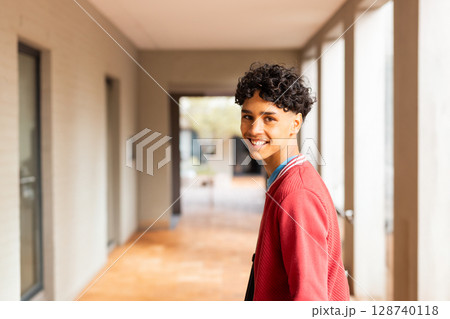 Teenage boy standing and looking back with smile wearing red jacket in campus corridor, copy space 128740118