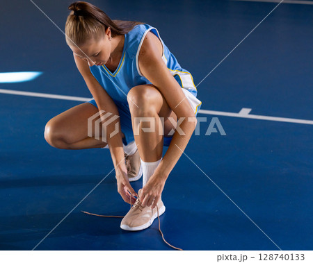 Female athlete tying sneaker laces on indoor court, wearing pastel sneakers and blue jersey 128740133