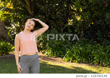 Asian woman performing neck stretch in backyard garden, wearing peach workout top and gray leggings Asian woman performing neck stretch in backyard garden, wearing peach workout top and gray leggings 128740148